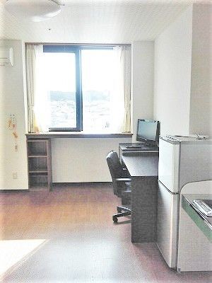 テレビが置かれたデスクや冷蔵庫、収納棚やキッチンがあるフローリングの居室。