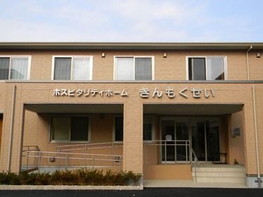 ２階建てのブラウン建物は玄関に階段と手すり付きのスロープがあり、背の低い木もたくさん植えられている。