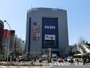 大きな商業ビルが建っていて、その前には横断歩道がある、様々な店舗の名前が外壁に書かれている。