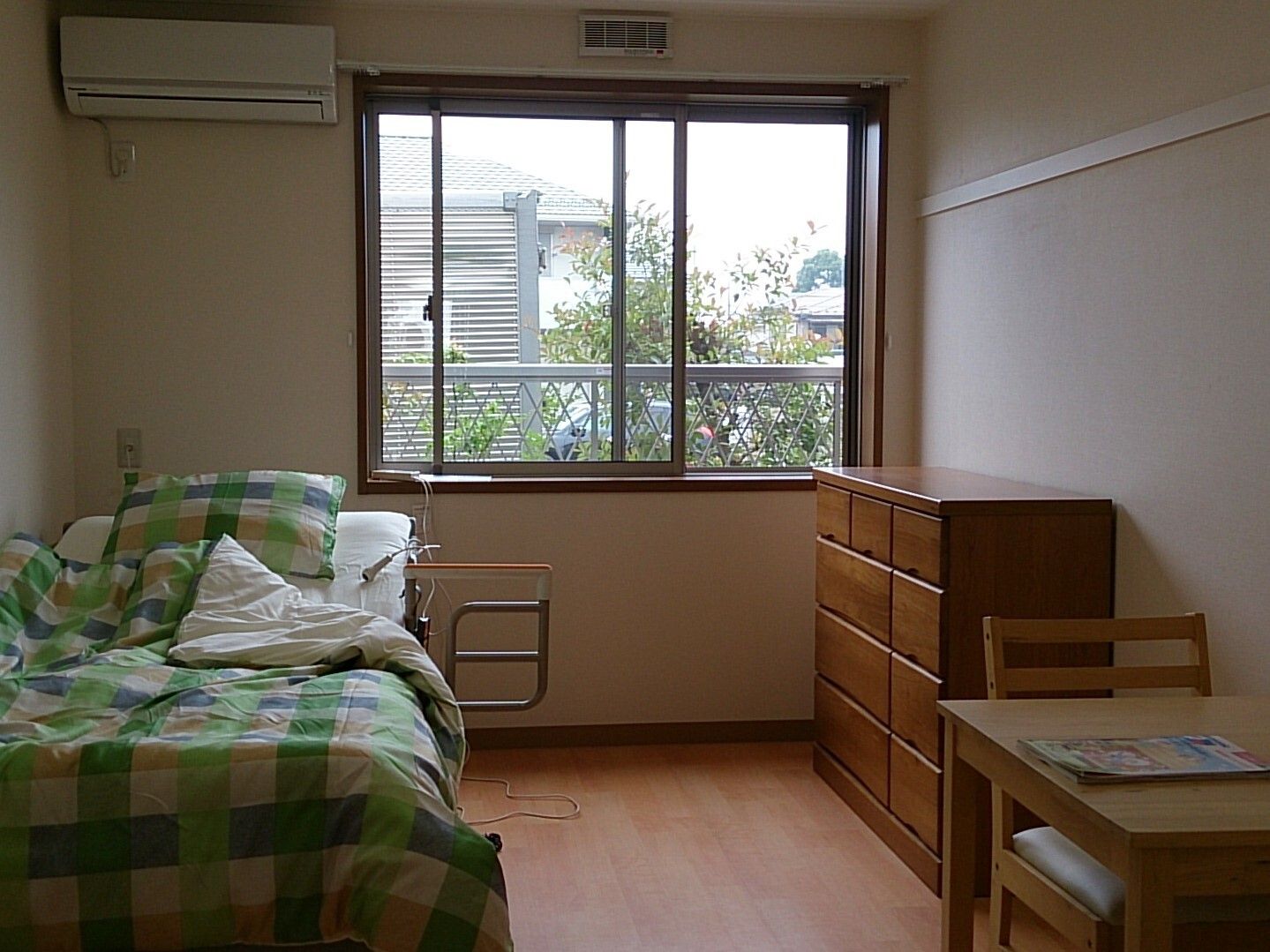 大きな窓が付いた部屋には寝具付きのベッドが置かれ、木製のタンスもある。フローリングのフロア。