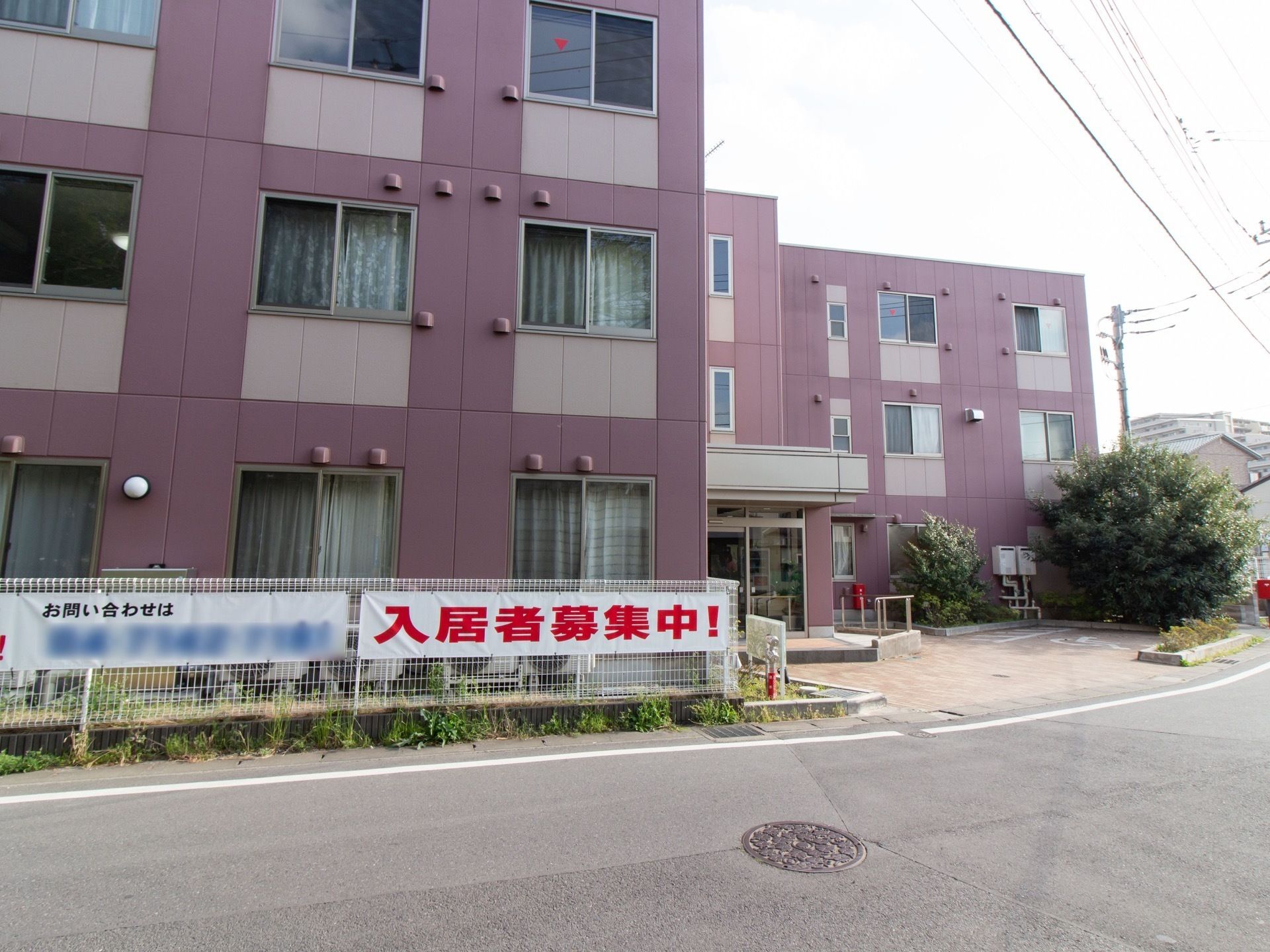 紫系統の色を基調とした３階建ての施設である。道路に面したフェンスに入居者募集中の横断幕が掲示されている。