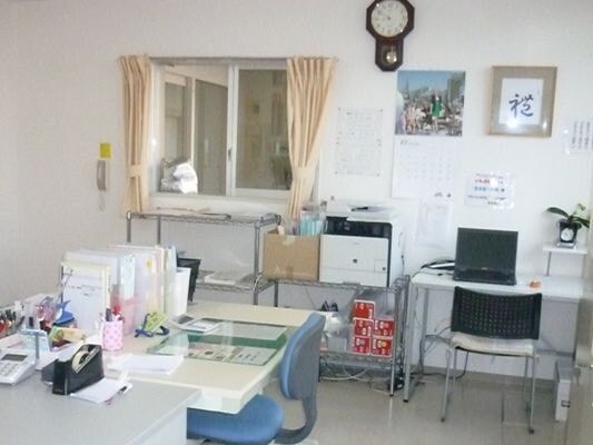 デスクが複数あり、書類やパソコンが置かれている。左側には受付けの窓もあり、壁にはカレンダーなどがある。