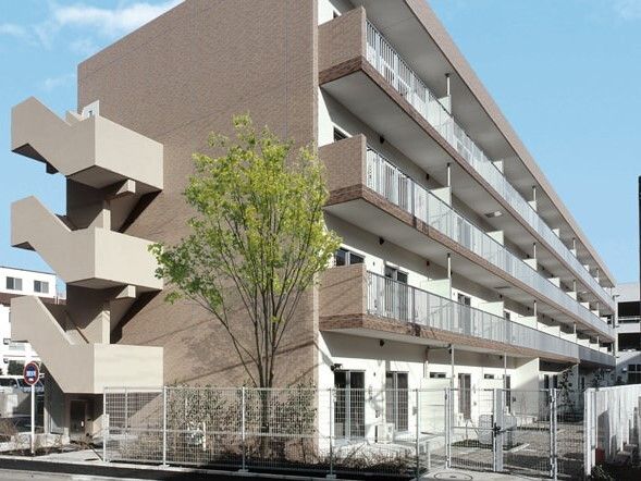 各フロアにベランダが付いたブラウン系の外壁の４階建ての建物。周りには草木が植えられ、フェンスもある。