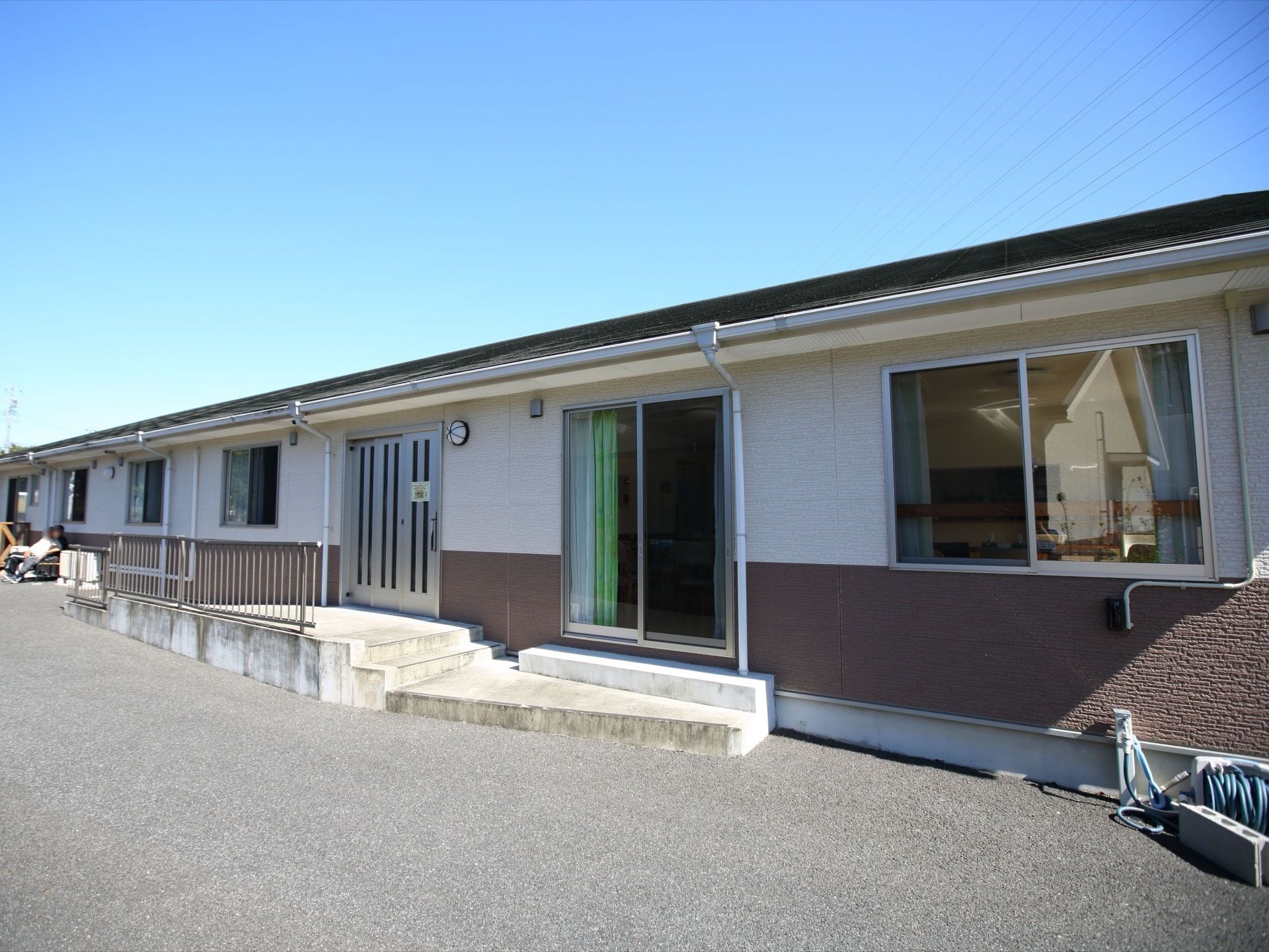 雲ひとつない晴天の日の平屋建て施設の外観である。入口には、3枚スライド式の扉と照明器具が設置されている。