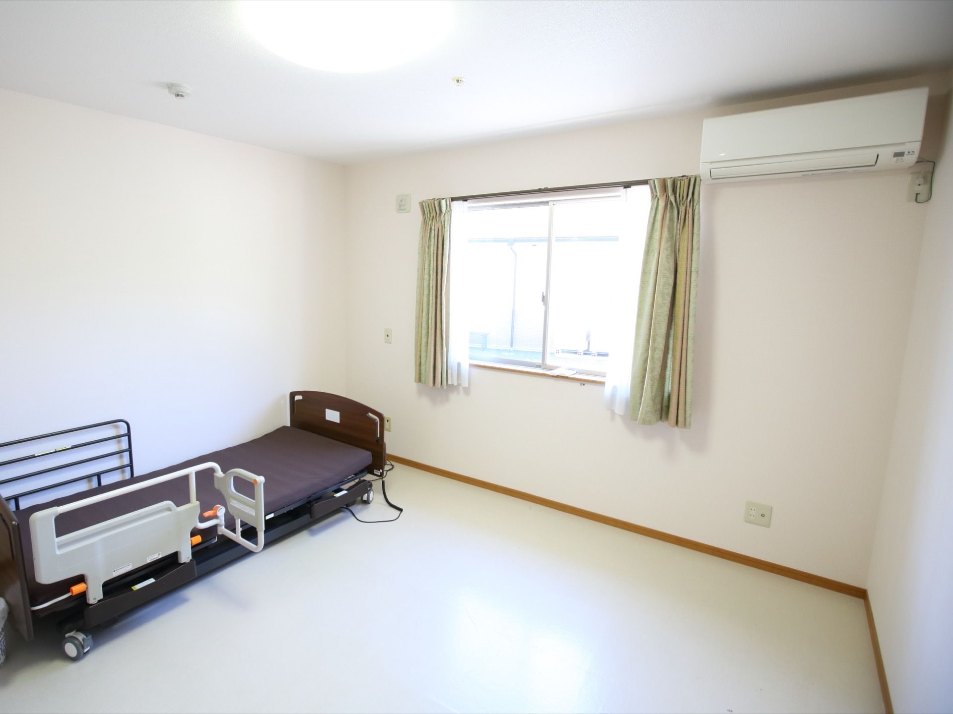 ご入居前の居室である。白い壁に窓やエアコンが設置され、介助バーが装着された介護用ベッドがある。