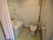 洗面台が備え付けられているトイレで、入口はピンク色のカーテンが取り付けられている。便器の前には、手すりの棒が設置されている。