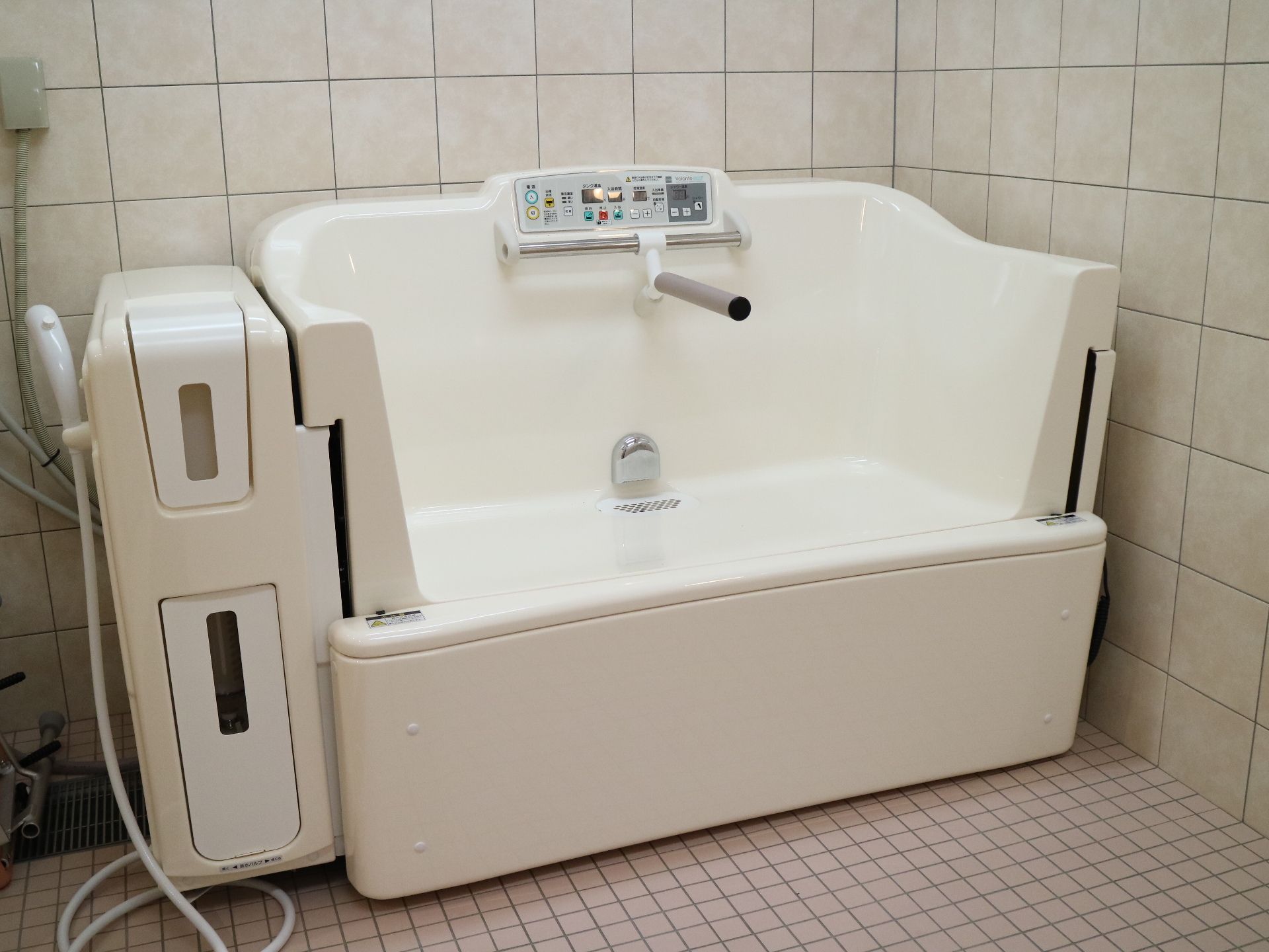 白い機械浴槽が設置された広い浴室はタイル張り。シャワーの前にはシャワーチェアもある。