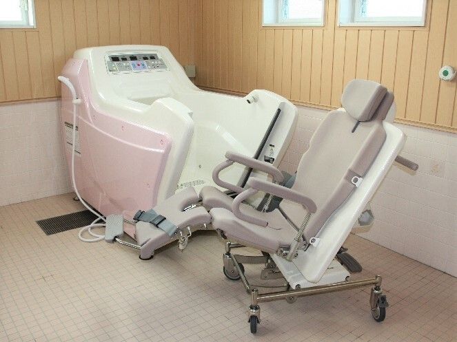 白とピンクの機械浴が設置されたタイル張りの浴室。車いすのような椅子部分が浴槽の壁となっている。機械にはシャワーも付いている。