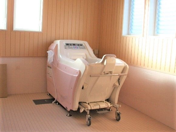 やさしい陽の光が入ってくる温かい雰囲気の浴室はタイル張り。白とピンクの座ったまま入浴可能な機械浴が設置されている。