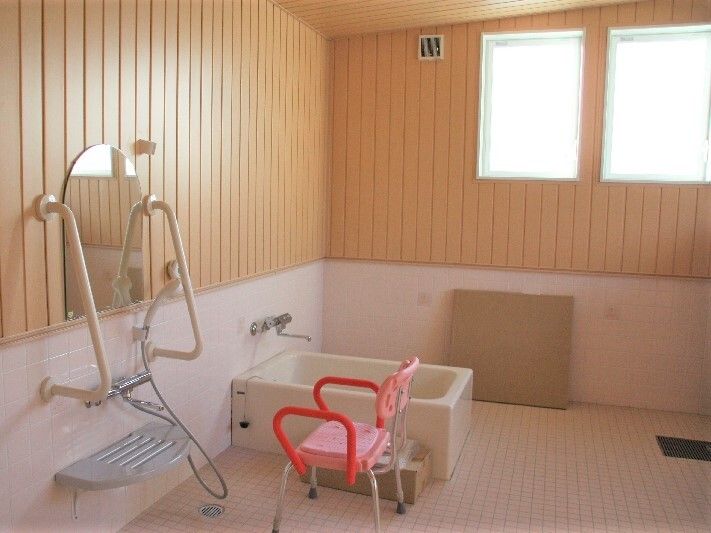 赤とピンクのシャワーチェアがシャワーの前に置かれ、３方向から介助ができる浴槽が設置され、シャワーの前の鏡の両サイドに手すりがある。