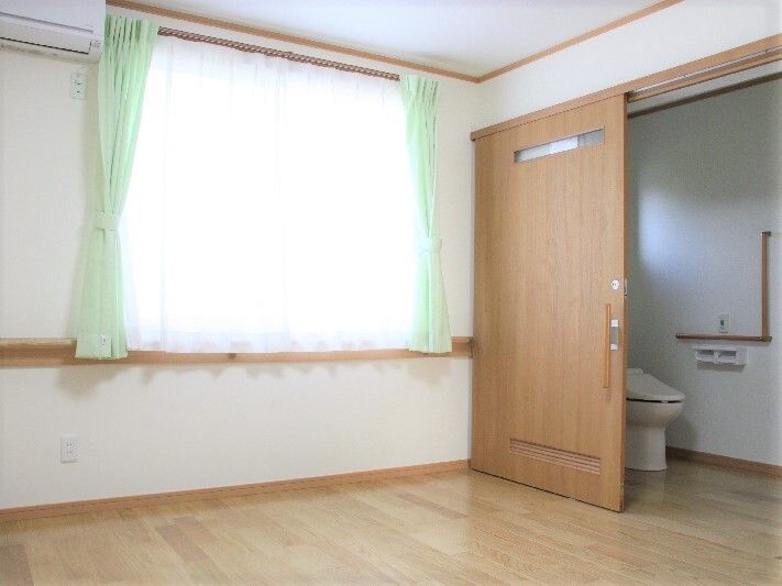大きな窓が付いた居室にはトイレも付いていて、トイレにはエル字型の手すりやトイレットペーパーホルダーがあり、スライド式のドアが設置されている。