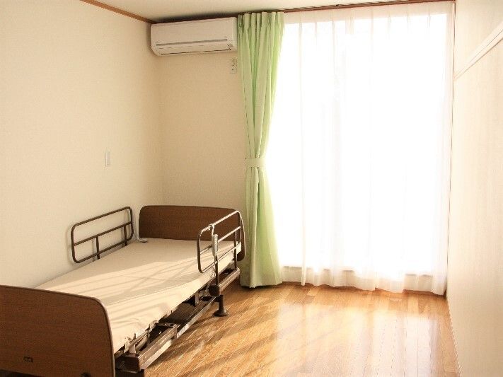 ピクチャーレールが付いたフローリングの部屋は大きな窓が付き明るい。カーテンやエアコン、介護用ベッドも設置されている。