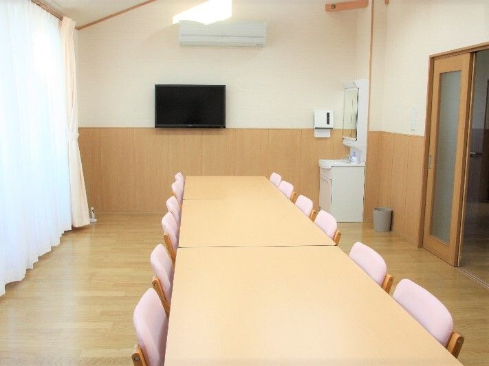 ピンクの椅子がセットされたダイニングテーブルがたてに二つ並べられ、大きな窓にはカーテンが付き、壁にはテレビが設置されている。