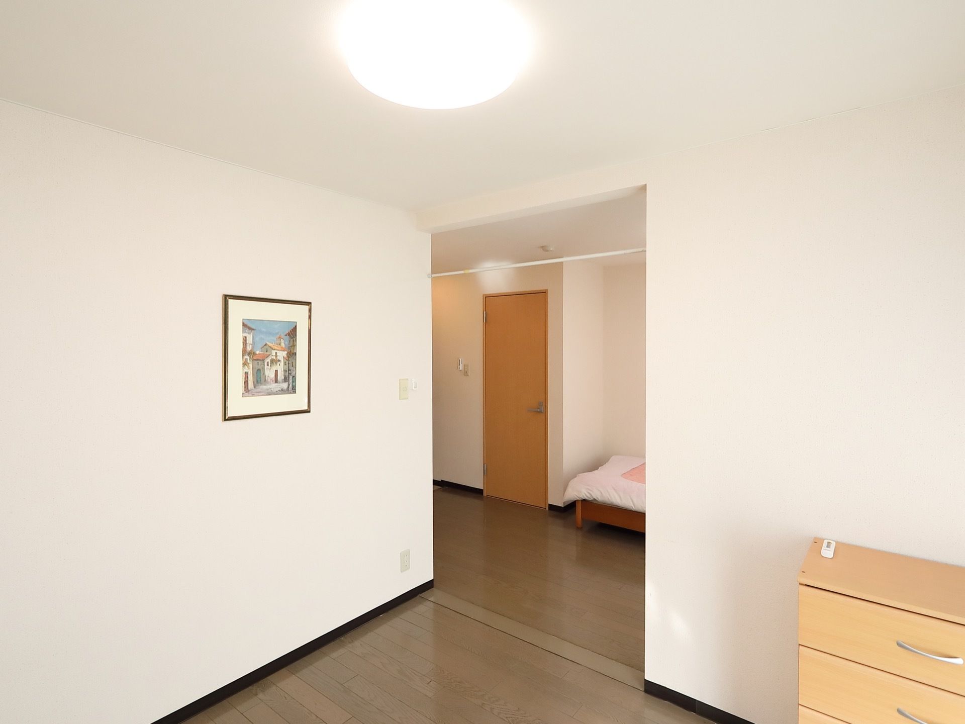 部屋数のある居室で、ベッドのある部屋と木製の小さなタンスが置いてあるスペースが別々である。タンスの上には、リモコンが置いてある。