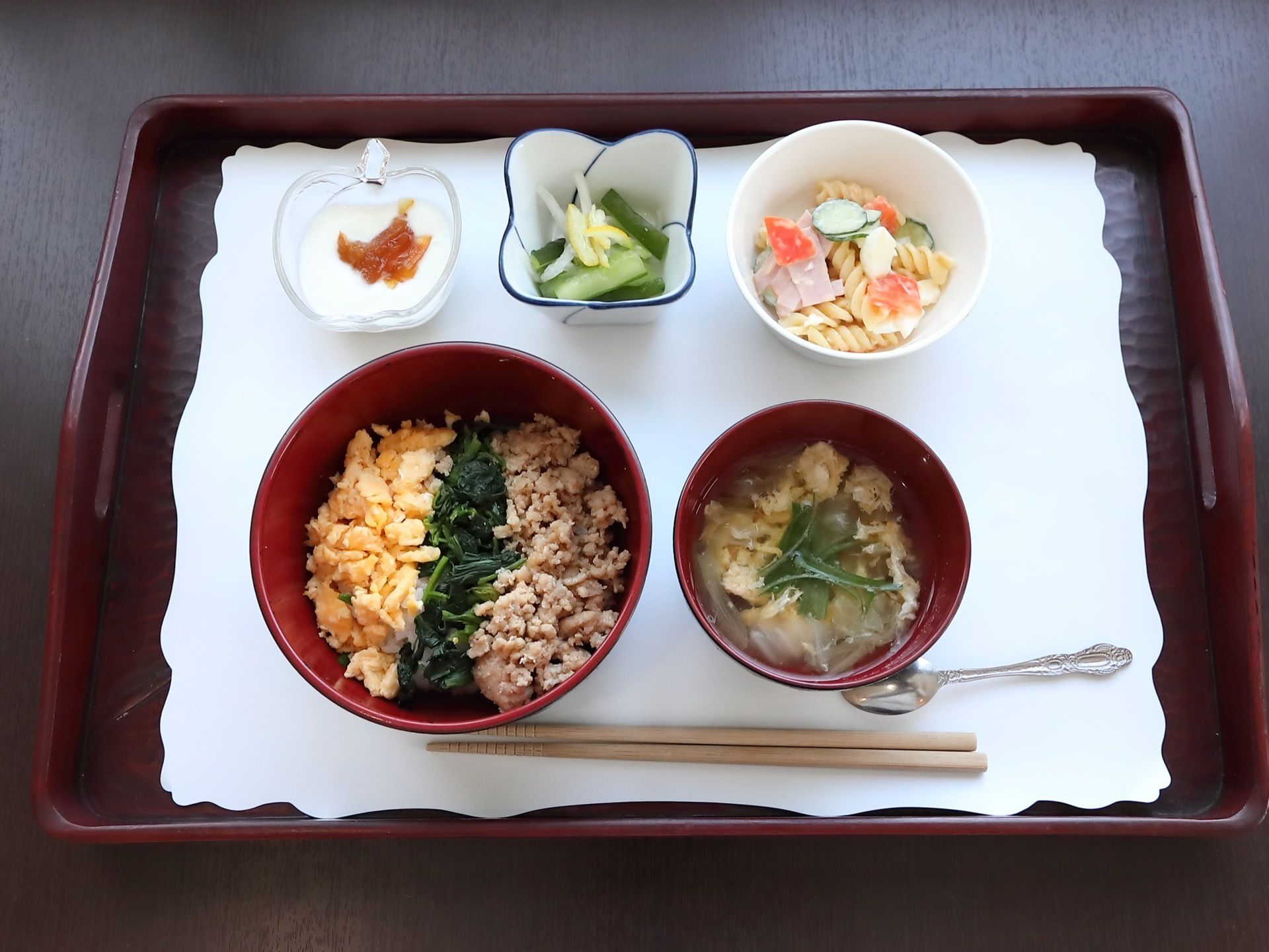 お盆の上に白い紙が敷かれ、三食丼、汁物、サラダ、漬物、ヨーグルトが置いてある。お箸とスプーンが用意されている。