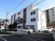４階建ての建物で、白い壁に部分的に茶色が使われたデザインである。建物の前と横には車が複数停まっている。