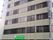 ベージュの９階建ての建物は鉄骨造りでどっしりとした印象。窓が多く、縦に長く、建物の周りには木も植えられている。