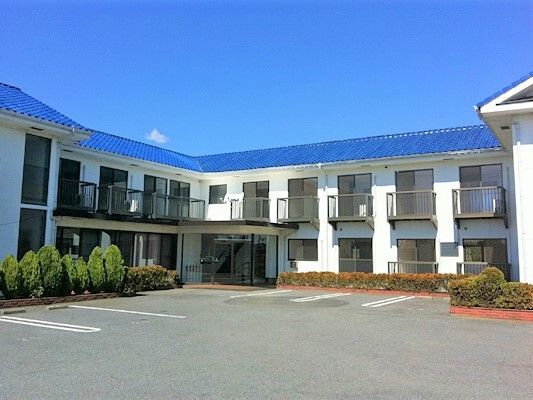 駐車場が付いた広い敷地内には2階建ての建物が立ち、玄関部分には屋根が付き、花壇には背の低い木が植えられている。