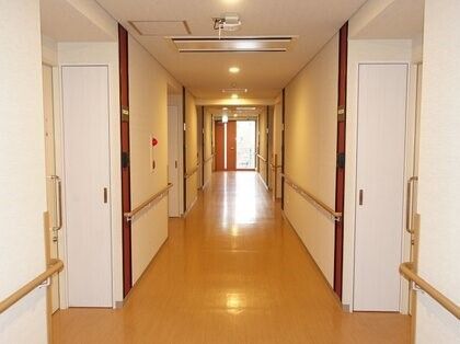 突き当りにエレベーターが完備された廊下である。白い壁には、明るい木目調の手すりや非常ベルが設置されている。