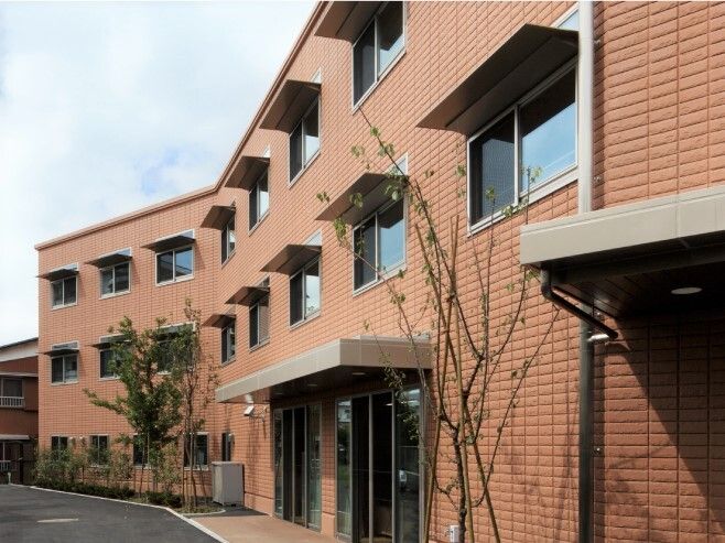 3階建ての建物はブラウン系でレンガ調の外壁。玄関には屋根が付き、各居室の窓には日差し除けが付いている。建物の周辺には大小の木も植えられている。