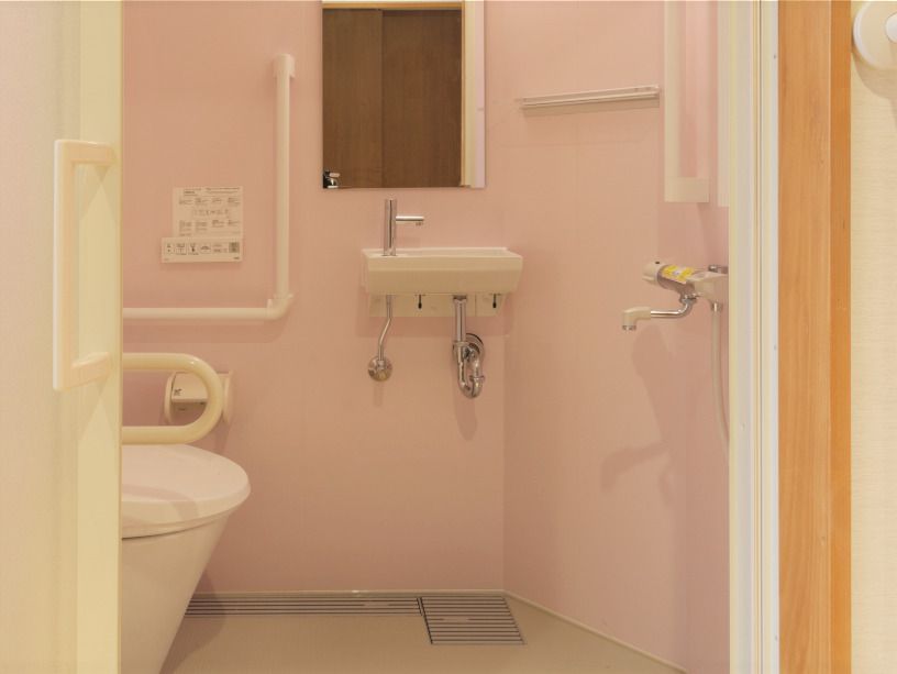 エル字型の白い手すりがピンクの壁に付いていて、スライド式のドアがあり、トイレ内に鏡が設置された手洗い場も付いている。