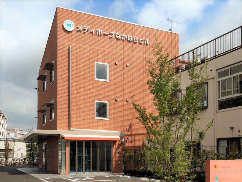 ブラウン系の3階建ての建物の外壁には住宅名が書かれ、敷地内には木も植えられている。玄関部分には屋根が付き、駐車スペースもある。