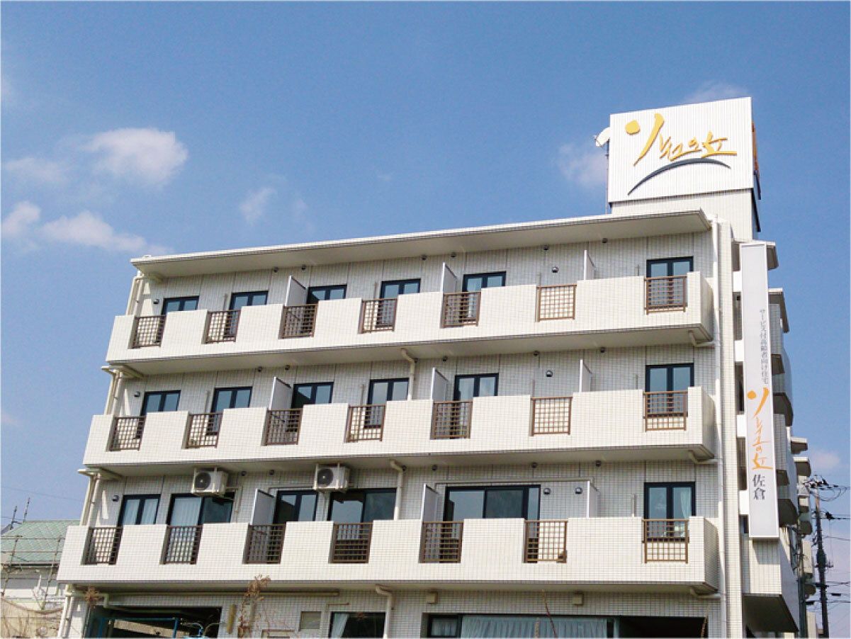 白い建物は4階建てで、２階以上のフロアにはベランダが付いている。住宅名が書かれた看板も付いている。