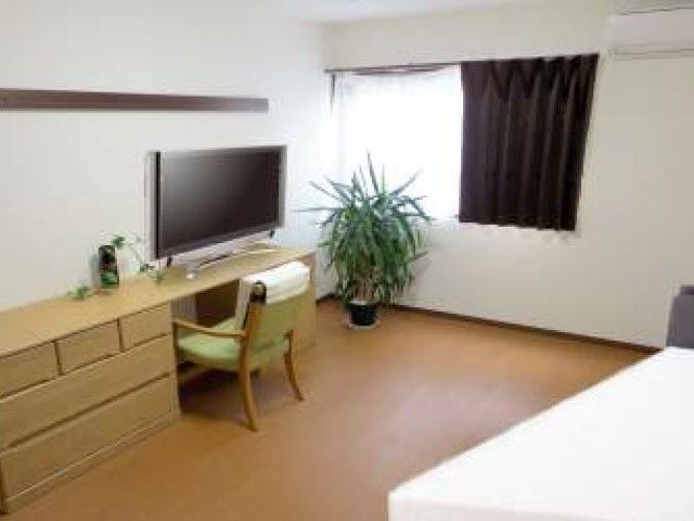 テレビとテーブル、たんすなどが並んでいる施設内の一室。広めのワンルームタイプの部屋の様子