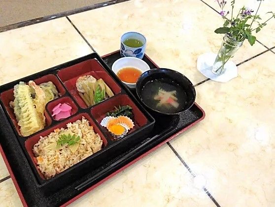 仕切りのついたお弁当箱に詰められた天ぷらや炊き込みご飯。その横には吸い物や天つゆ、お茶もある。