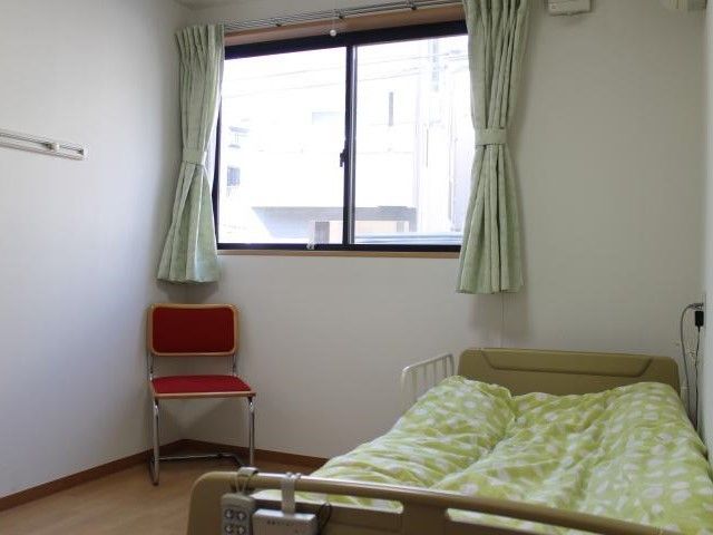 居室には淡いグリーンのカーテンが付いた窓があり、明るい光が差し込む。ベッド横には椅子も置かれている。
