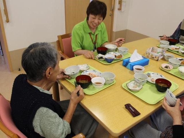 食事風景。ダイニングルームに入居者が集まり、スタッフと食卓を囲み、家庭的な雰囲気で食事の時間を過ごしている。