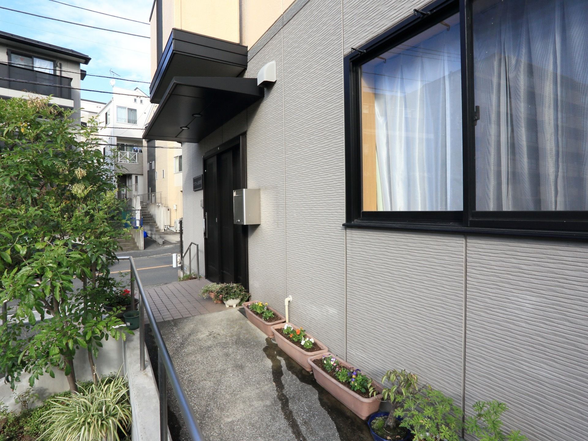 玄関まではスロープと階段がある。建物に沿ってプランターに植えられた花が飾られている。入口横にポストが設置されている。