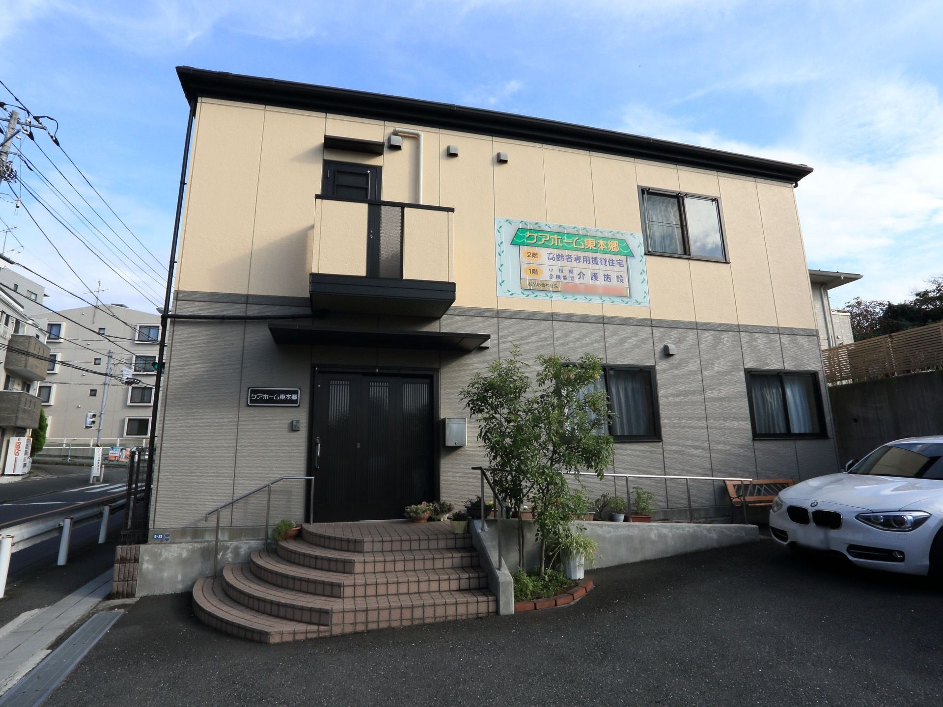 ベージュとグレーに塗り分けられた外観で、玄関前に扇状に階段が広がっている。施設前は駐車スペースになっている。