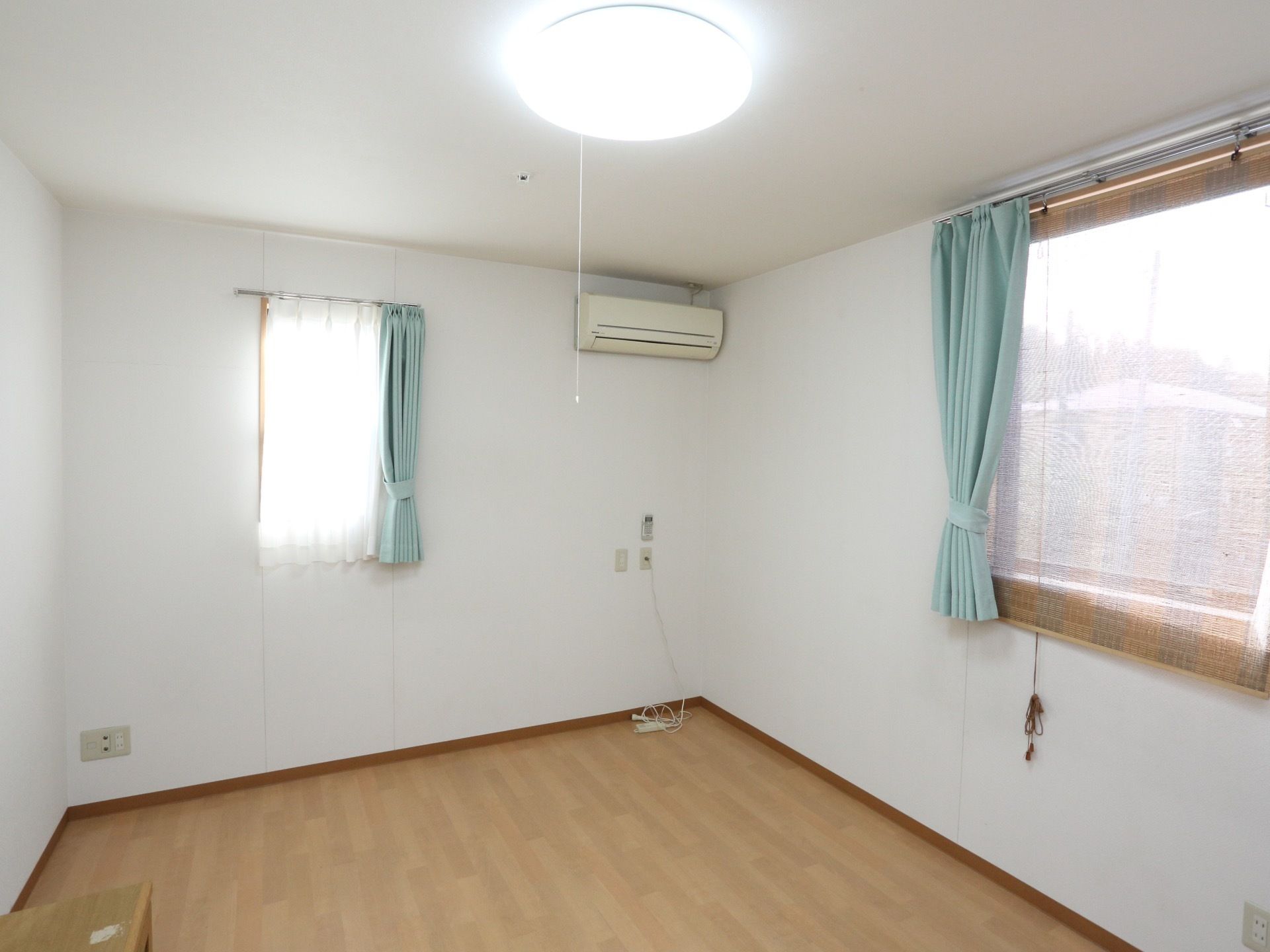角部屋には窓が２か所に開いている。水色のカーテンが掛けられ束ねられている。天井近くにエアコンが設置されている。