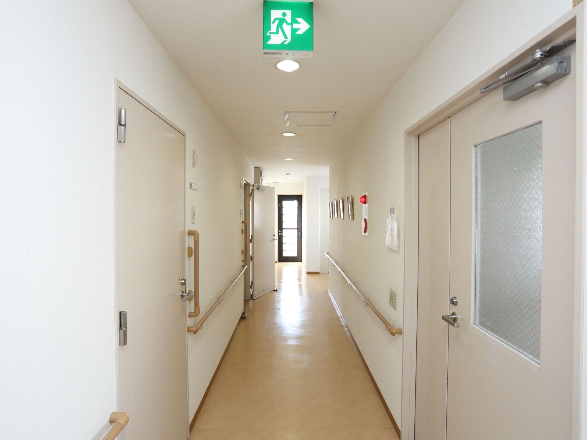 廊下の両側に手すりが設置されている。天井に非常口サインが表示され、壁には非常ベルのボタンがある。ダウンライトで照らされている。