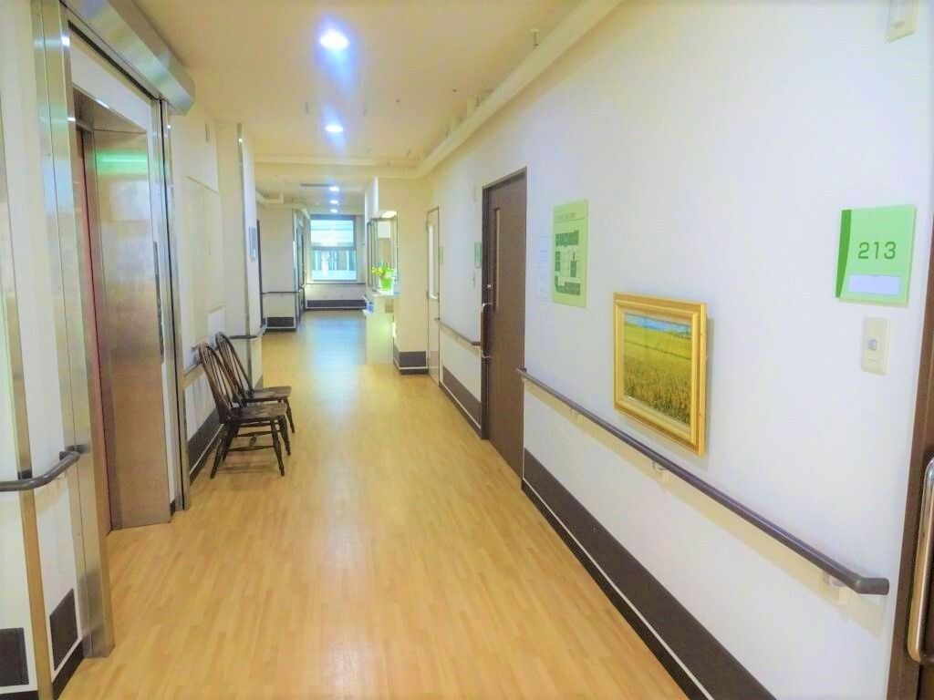 スライド式のドアが設置された居室が右側に並び、左側にはエレベーターと数脚の椅子があり、廊下はまっすぐ続いている。