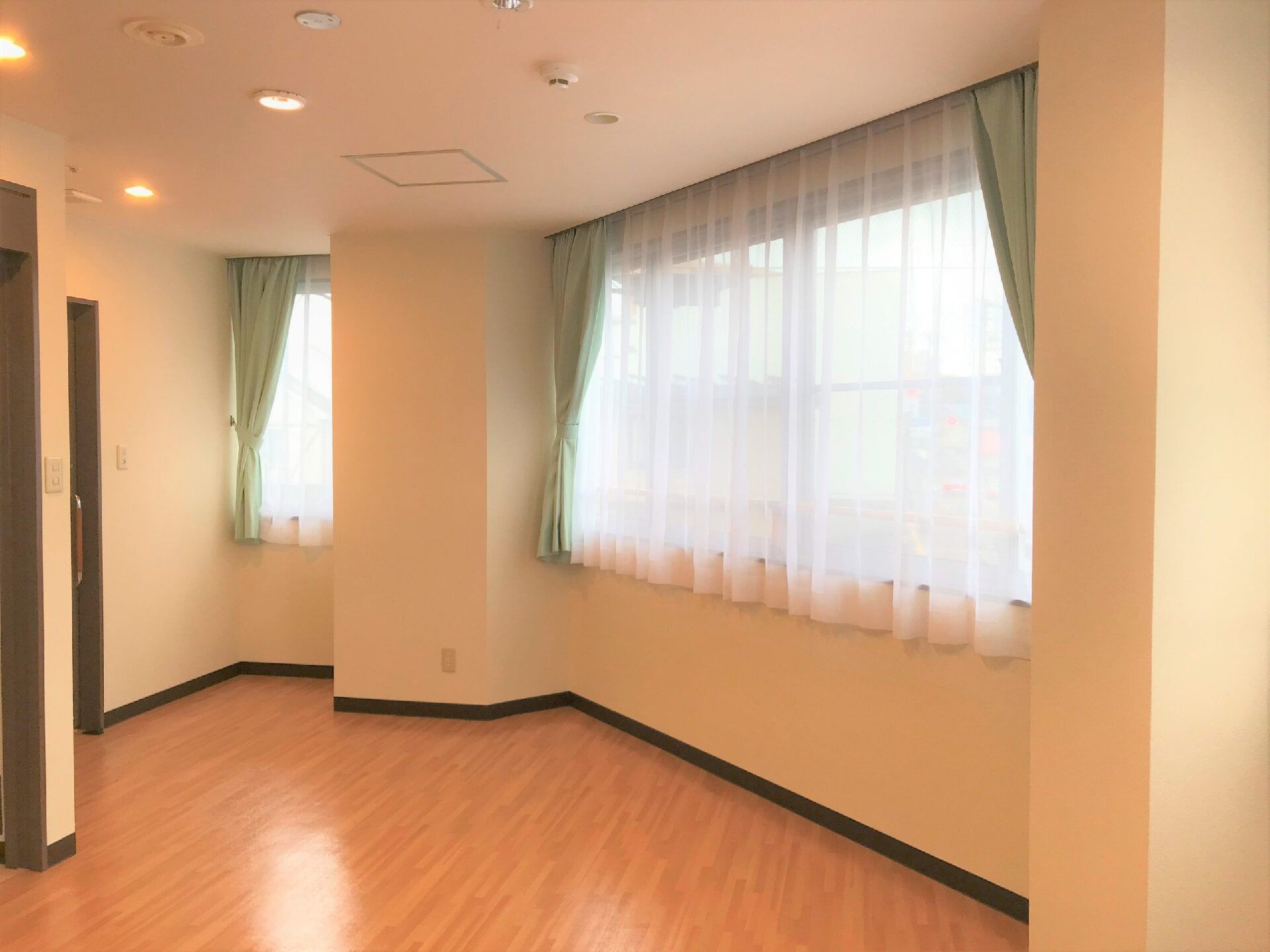 大小のカーテン付きの窓があるフローリングの居室。天井にはスプリンクラーが付き、家具などは置かれていない。日当たり良好。