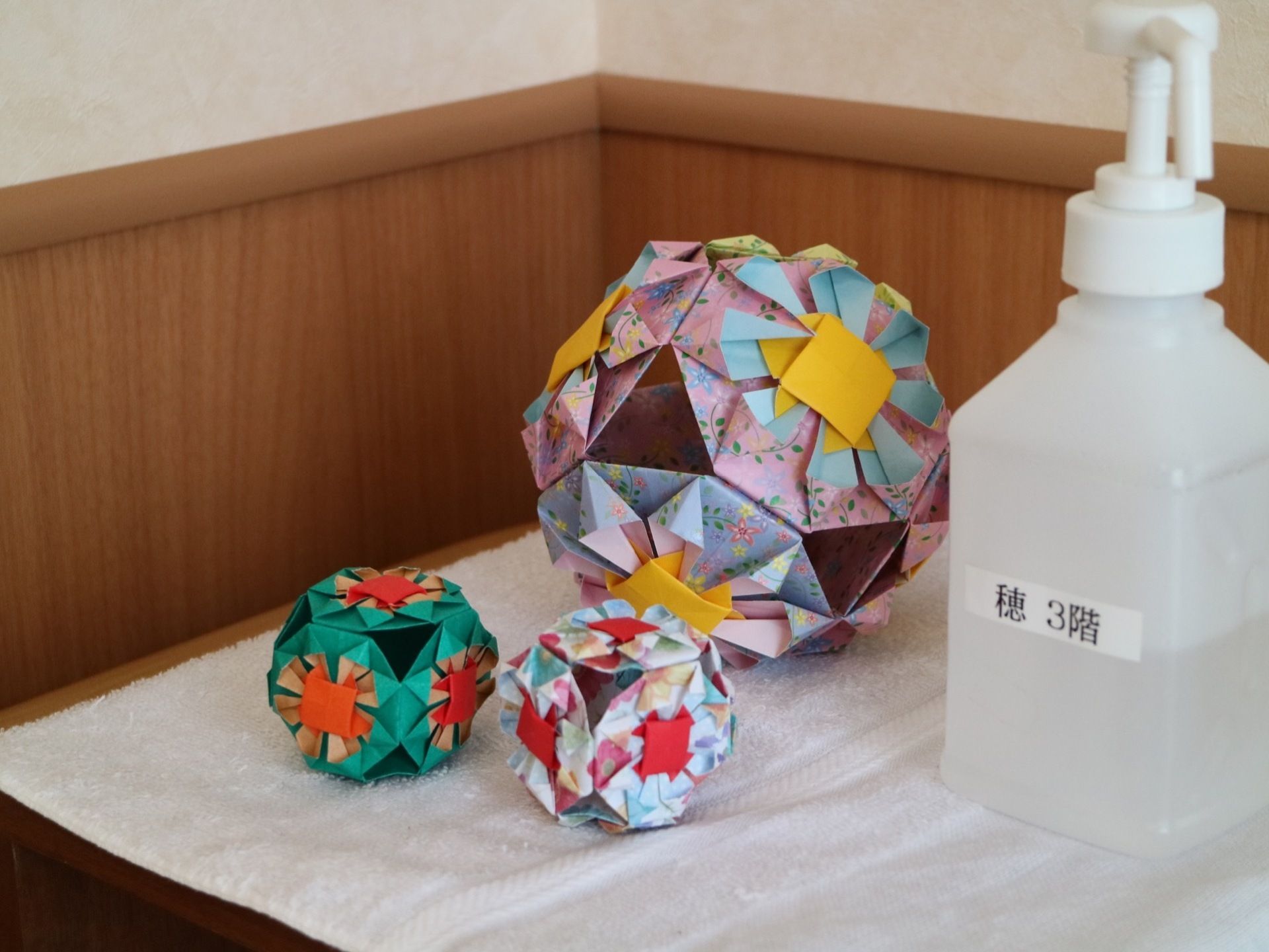木製の棚の上に、折り紙で作られた装飾とアルコール消毒液が置かれている。下には白いタオルが敷かれている。