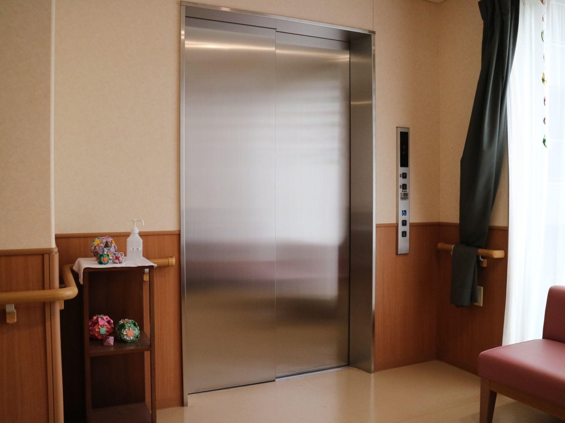 エレベーター横に木製の台が設置されており、上に消毒液が置かれている。行先ボタンは、車椅子をご利用の方も押しやすいよう配慮されている。