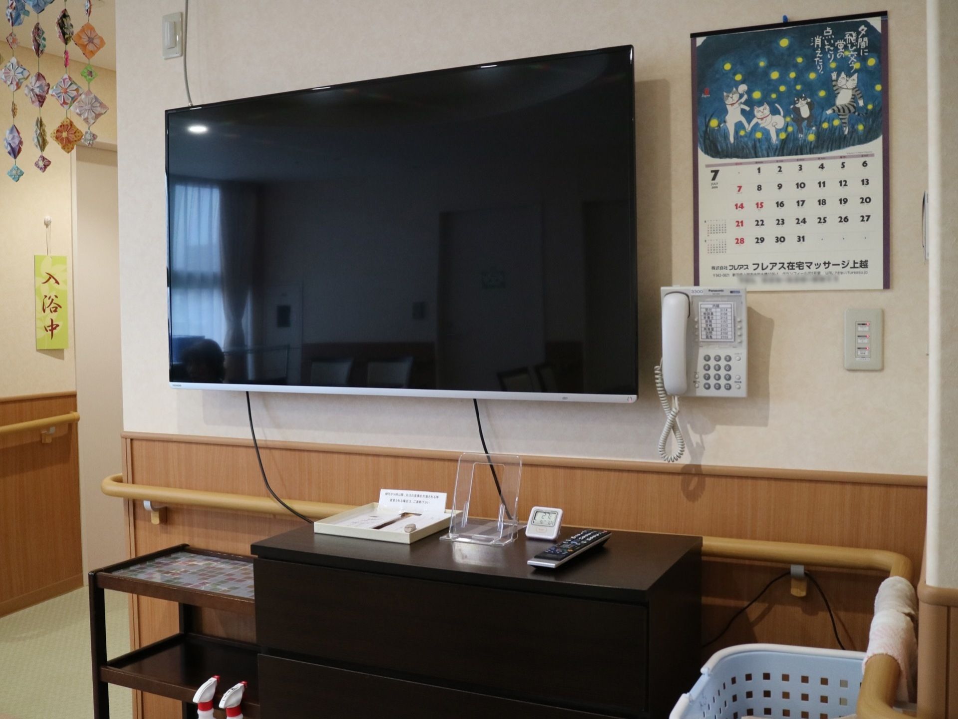 食堂の壁に、テレビや電話、手すりが取り付けられている。下に設置されている収納棚の上には、リモコンが置かれている。