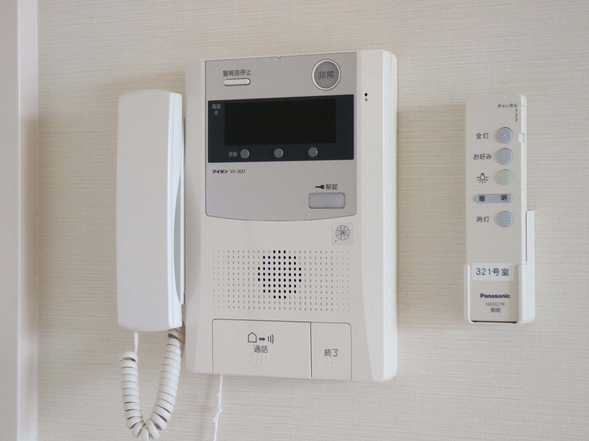 部屋の壁にインターホンが設置されており、受話器で話せるようになっている。横には、照明のリモコンが取り付けられている。