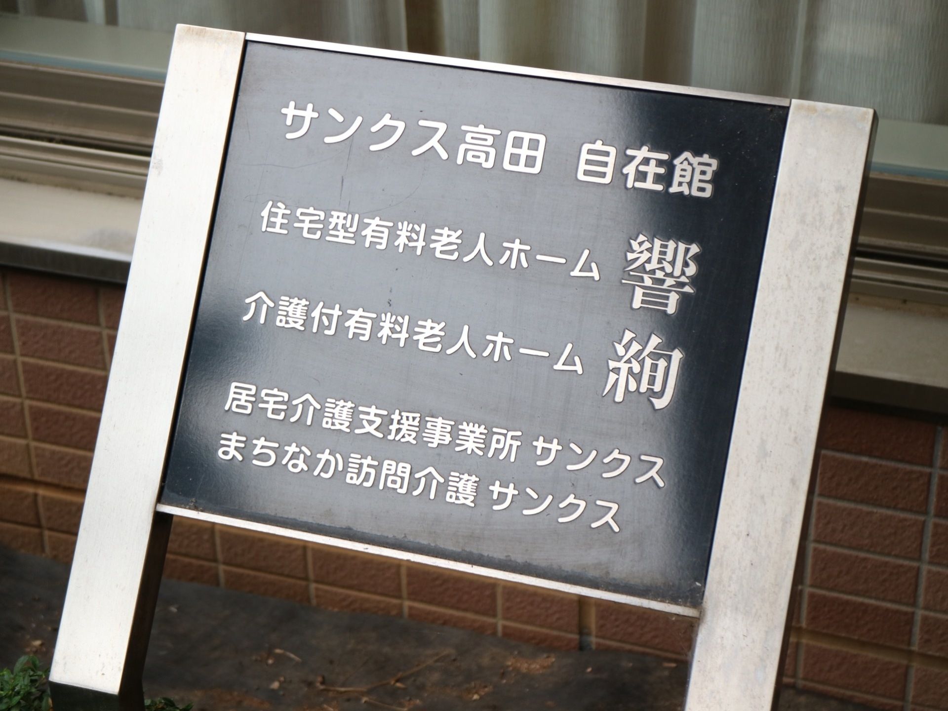 施設前に看板が設置されており、黒いベースにシルバーで施設名が書かれている。外壁はタイル調となっている。