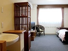 車いす対応の洗面所が付いたカーペット敷きの部屋には寝具付きのベッドやいす、収納家具が置かれている。