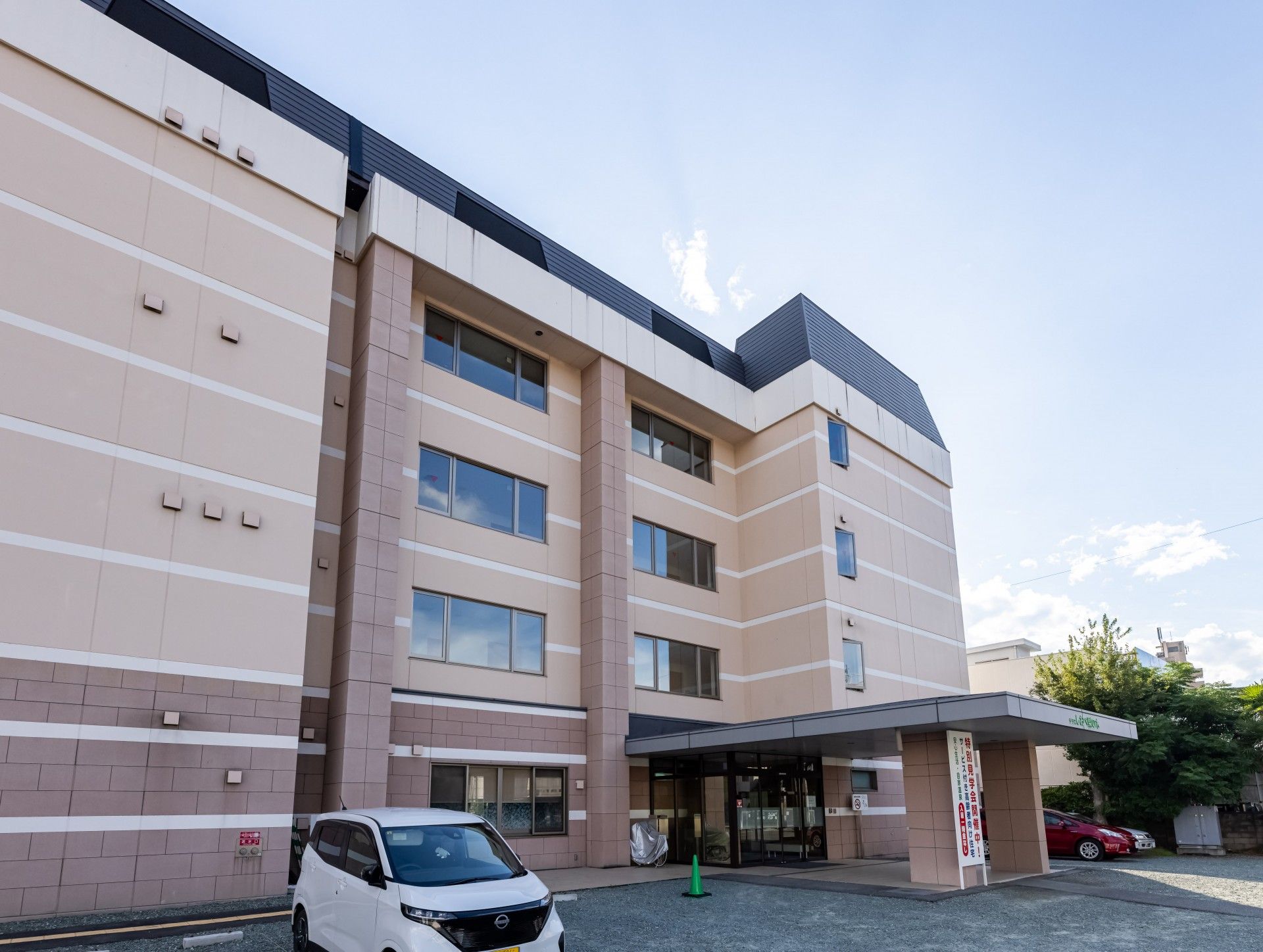 白いラインが入ったブラウン系の建物。3階建てで屋外階段が設置され、玄関には車寄せが付き、自動ドアもある。