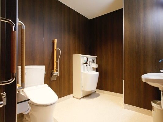 ダークブラウンの壁のトイレは広く、可動式の手すりと固定式の手すりが付き、手洗い場もある。