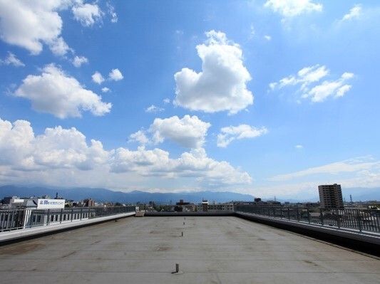 白い雲が点在している青い空の下に広がる街並みを眺めることができる屋上。ベンチなどは置かれていない。