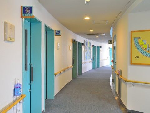 扇型の建物に沿って緩やかにカーブした廊下の両側に部屋のドアが並んでいる。壁には手すりが設置されており、手すりの上に消毒液が置かれている。