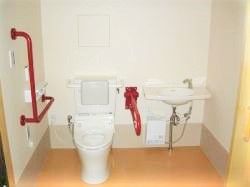 L字型の赤い手すりが付いた広めのトイレの様子。横に洗面台が付いている
