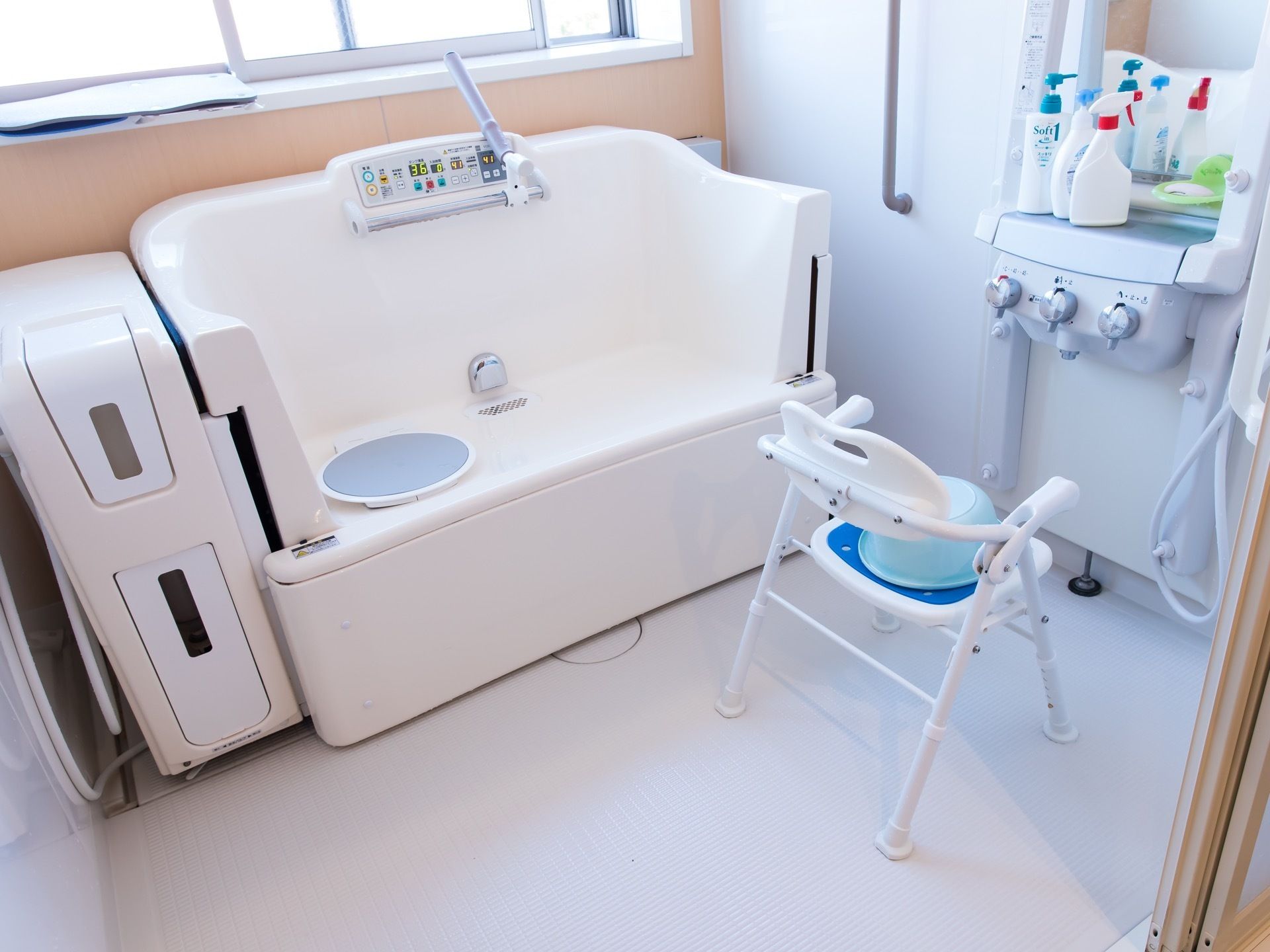 窓際に機械浴用の浴槽が設置されている。洗い場には浴用チェアーが置かれている。壁際の鏡の下の棚部分にシャンプーなどがある。