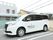 駐車場に白い３列シートのミニバンが停まっている。車体には運営会社名が印字されている。奥には住宅街が見える。